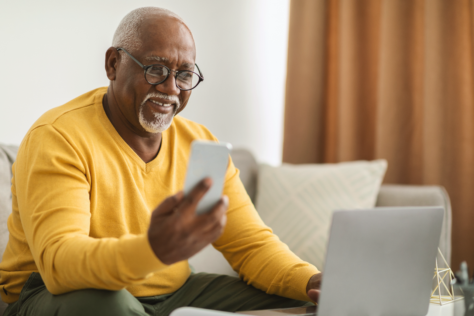 A smiling man on a couch looks at his phone while working on a laptop.